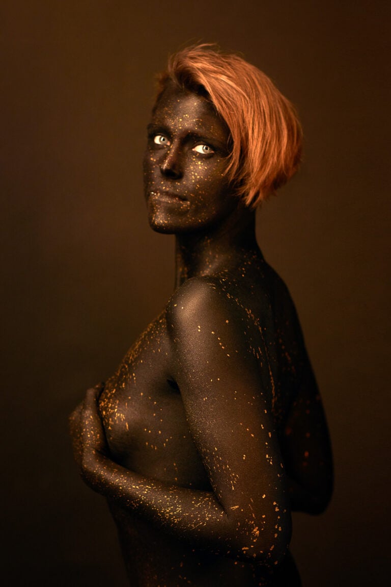 Bodypainting-Porträt einer Frau mit schwarzer Körperfarbe und goldenen Farbsprenkeln vor warmem braunem Hintergrund