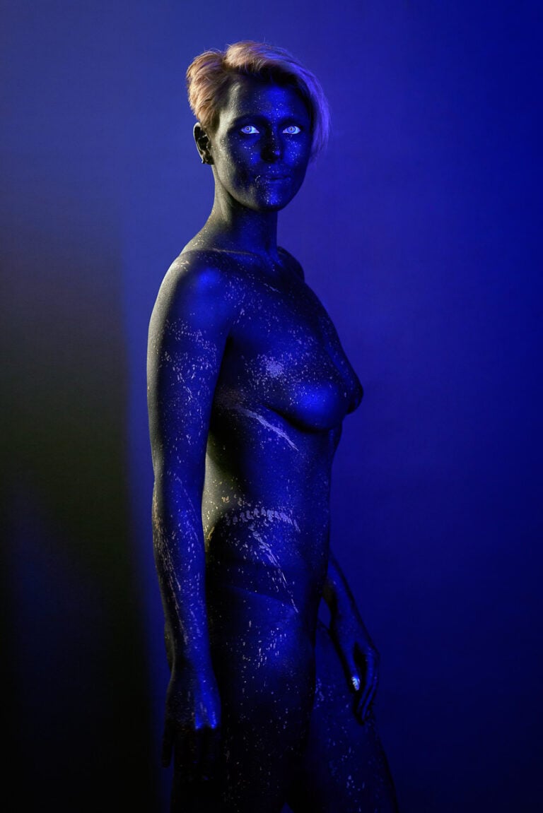 Bodypainting-Aktporträt in Blau mit leuchtenden Augen und Farbsprenkeln vor dunklem Studiohintergrund