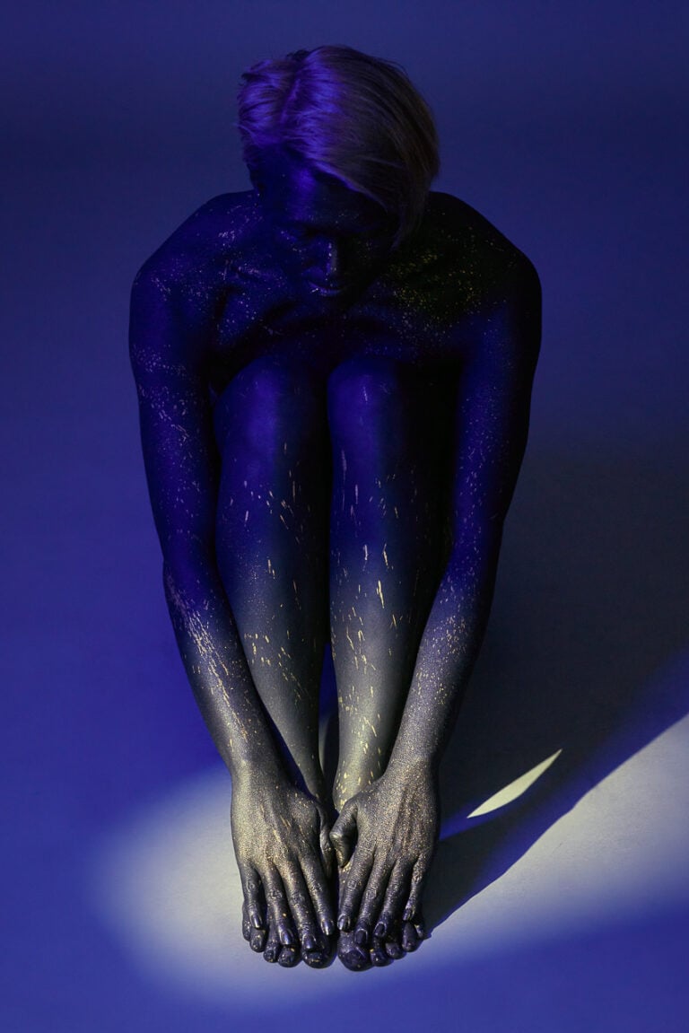 Bodypainting-Akt in sitzender Pose mit blauer Farbe und goldenen Sprenkeln im Studio