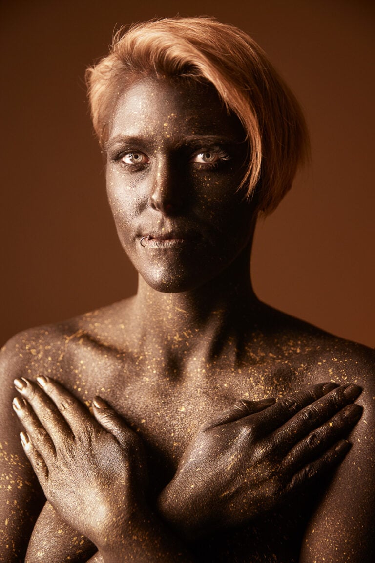 Bodypainting-Porträt in Gold und Schwarz mit gekreuzten Händen vor warmem Hintergrund