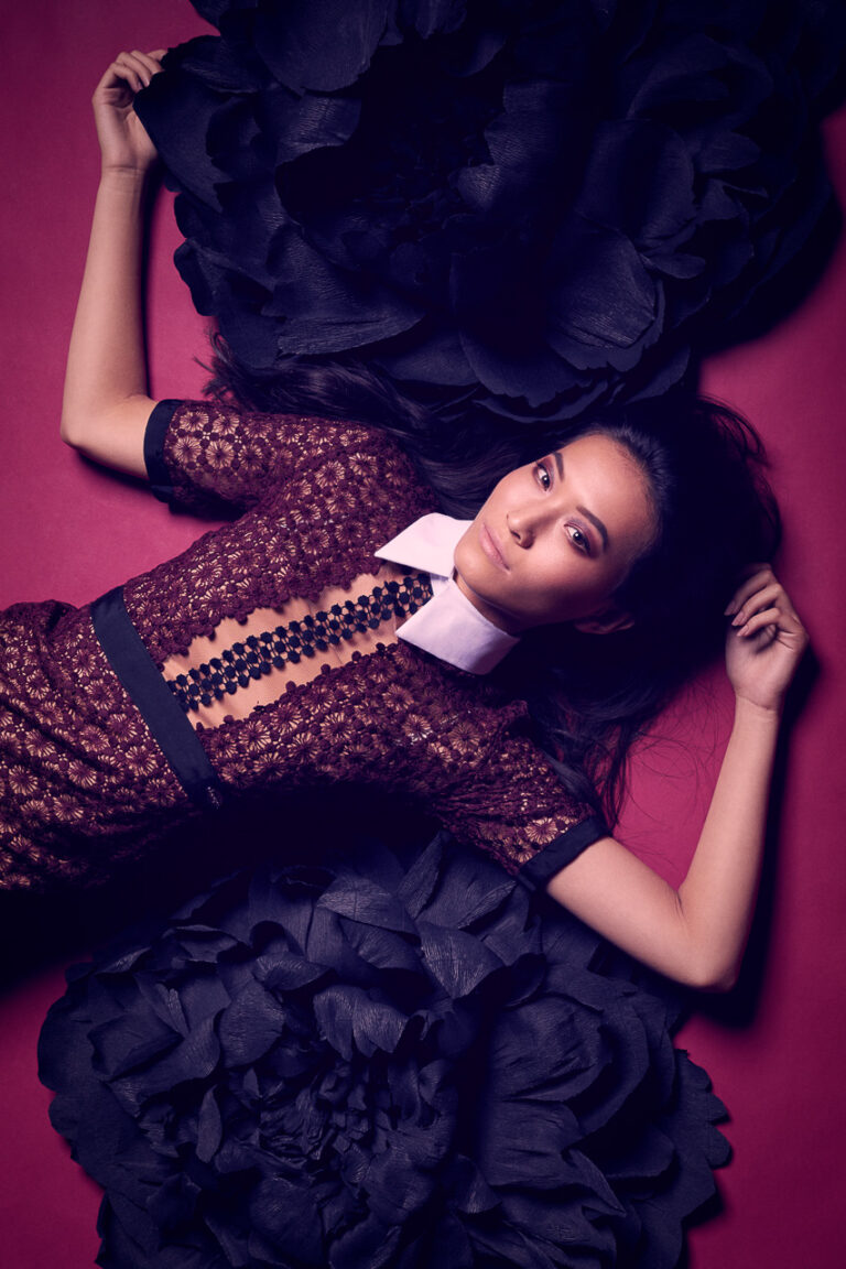 Fashion-Porträt einer Frau im dunklen Spitzenkleid zwischen großen schwarzen Blüten auf violettem Hintergrund