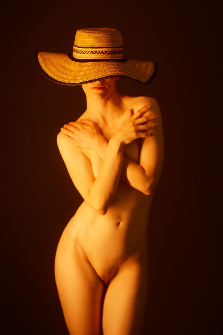 Aktportrait einer stehenden Frau mit großem Strohhut im warmen, goldenen Licht vor dunklem Hintergrund