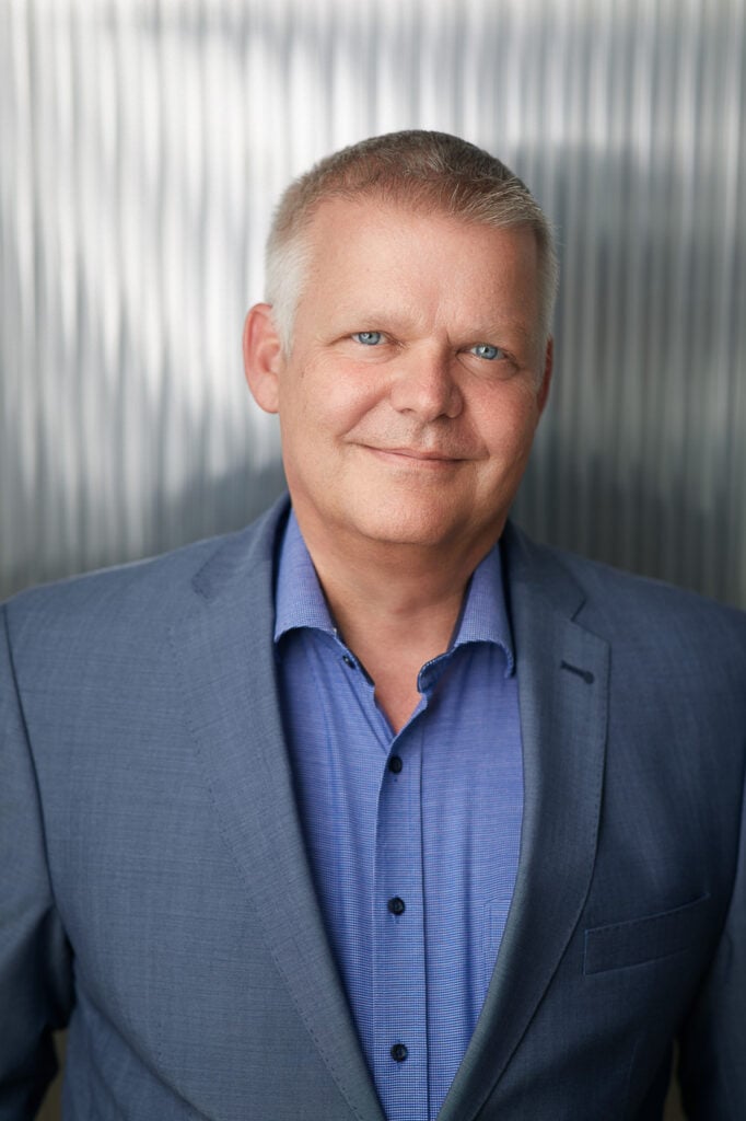 Geschäftsmann mit warmem Lächeln im Business Portrait vor strukturierter Wand – locker inszeniert für LinkedIn und Website.